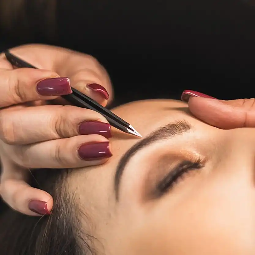 Close-up of a beauty therapist with burgundy nail polish using black tweezers to precisely shape and define a client's eyebrow at Unique Beauty & Style salon in Perth.