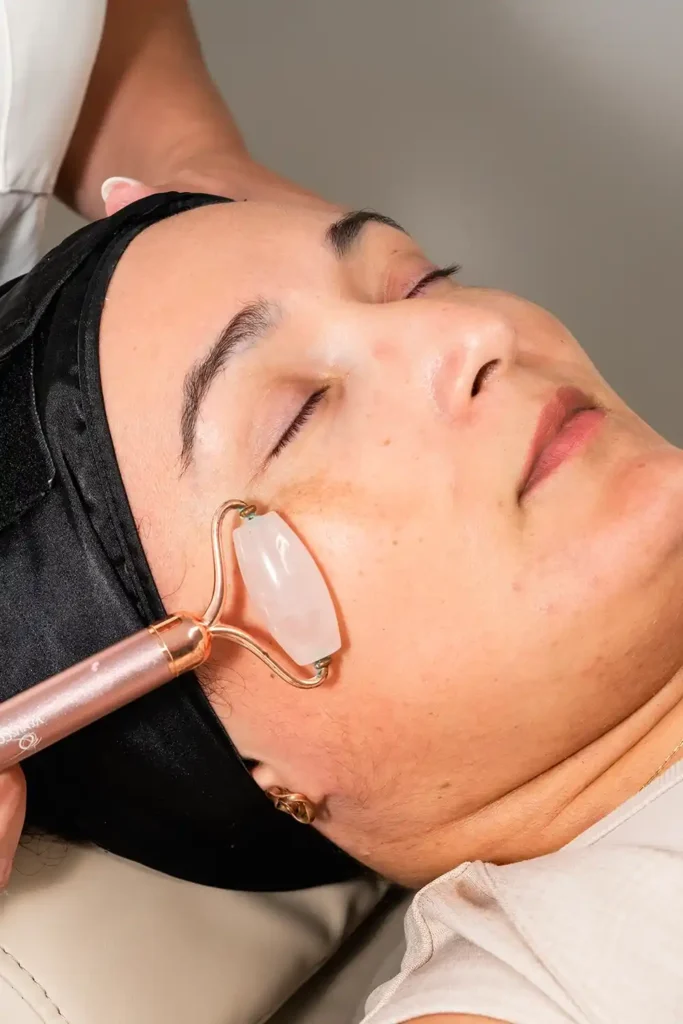 Close-up of a relaxing facial treatment using a rose quartz facial roller at Unique Beauty & Style salon in Perth. The client rests with her eyes closed, wearing a black spa headband, as the esthetician gently massages her cheek.