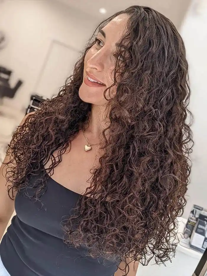 A smiling woman showcasing voluminous, long dark brown curly hair extensions at Unique Beauty & Style salon in Perth. The beautifully defined extensions blend seamlessly with her natural hair. She is wearing a black top and a delicate gold heart necklace.