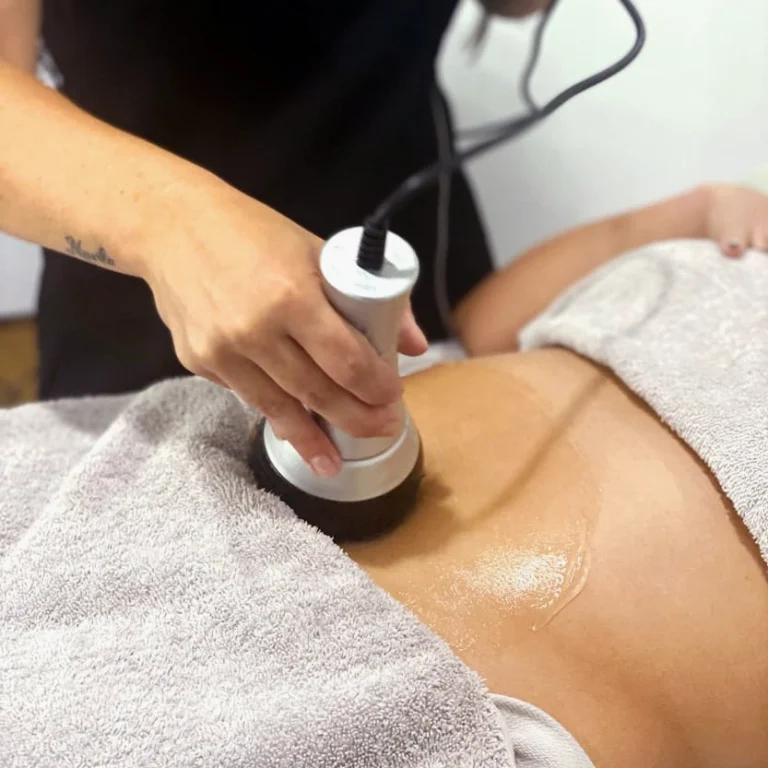 A professional aesthetician at Unique Beauty & Style in Perth, wearing a black uniform, black gloves, and a face mask, applies a creamy body treatment product onto the back of a client. The client is lying face-down on a massage table covered with white towels, with her eyes closed and hair tied up. The scene takes place in a bright, clean spa treatment room, with blurred product bottles on shelves visible in the background, emphasizing relaxation and skin care.