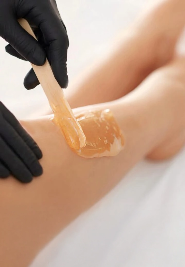 A detailed close-up of an aesthetician wearing black gloves using a wooden spatula to apply warm amber-colored wax to a client's lower leg at Unique Beauty & Style salon in Perth, Australia.
