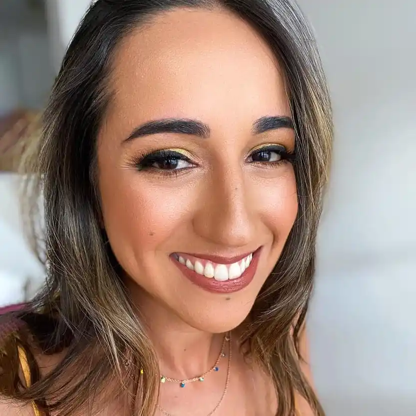 Close-up portrait of a smiling woman showcasing flawless professional makeup done at Unique Beauty & Style salon in Perth. Her beautiful makeup features a glowing complexion, perfectly defined eyebrows, golden eyeshadow with black eyeliner, and a warm nude lipstick. She is wearing a delicate gold necklace with small colorful beads.
